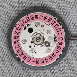 Puede incluir: Primer plano de un movimiento de reloj con una rueda de fecha rosa y engranajes plateados. La rueda de fecha muestra n&uacute;meros del 1 al 31. El movimiento del reloj est&aacute; sobre un fondo gris.