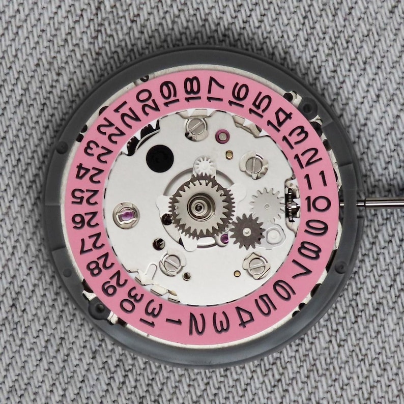 Puede incluir: Primer plano de un movimiento de reloj con una rueda de fecha rosa y engranajes plateados. La rueda de fecha muestra n&uacute;meros del 1 al 31. El movimiento del reloj est&aacute; sobre un fondo gris.