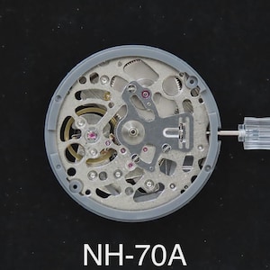 May include: A detailed view of a watch movement, featuring intricate gears, springs, and jewels. The movement is encased in a gray frame, with a clear tool inserted. The text "NH-70A" is displayed at the bottom.