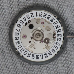 Puede incluir: Primer plano de un movimiento de reloj con una esfera blanca y un anillo exterior gris. La esfera muestra los números del 1 al 31, que indican los días del mes. Los intrincados engranajes y componentes internos son visibles.