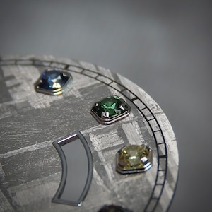 May include: Close-up of a watch face with a textured, gray surface. The face features three square-cut gemstones in blue, green, and yellow, each set in a silver bezel. A silver-colored, curved element is also visible.