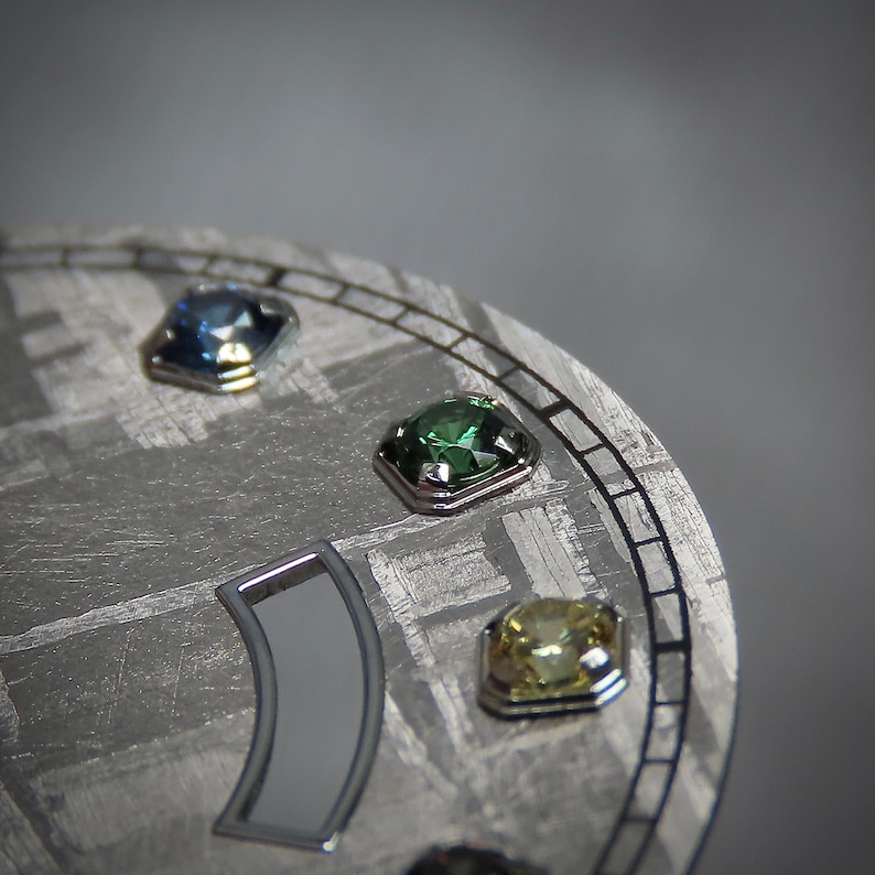 May include: Close-up of a watch face with a textured, gray surface. The face features three square-cut gemstones in blue, green, and yellow, each set in a silver bezel. A silver-colored, curved element is also visible.