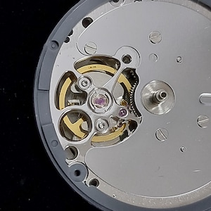 以下が含まれることがあります： 時計のムーブメントのクローズアップ。複雑な機械部品が写っています。銀色のベースには、金色の歯車、テンプ、小さな宝石が収められています。デザインは円形と曲線の要素を取り入れ、精密なエンジニアリングに焦点を当てています。