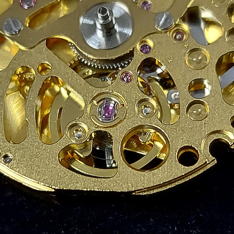 May include: Close-up of a gold watch mechanism. The intricate design features gears, screws, and jewels. The watch parts are arranged on a gold-toned base, showcasing the precision of the timepiece.
