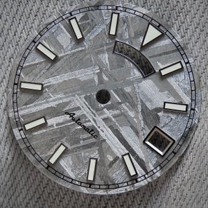 Puede incluir: Una esfera de reloj con un patrón gris texturizado único, posiblemente meteorito. Cuenta con marcadores de hora rectangulares blancos y un borde negro. La palabra "Automatic" está escrita en cursiva. Las manecillas y los marcadores tienen un acabado plateado.