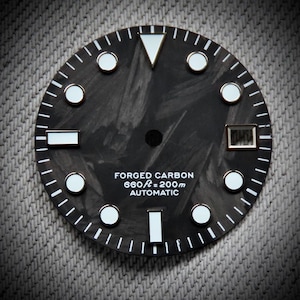 Puede incluir: Una esfera de reloj de carbono forjado negro con marcadores de hora y agujas blancos. La esfera tiene un patrón texturizado y el texto "FORGED CARBON 660ft = 200m AUTOMATIC". La esfera del reloj tiene una ventana de fecha a las 3 en punto.