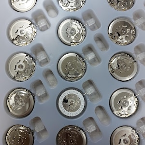 May include: A collection of watch movements, featuring intricate gears and components, arranged in a light gray plastic tray. The movements are silver-toned, showcasing the precision engineering of the watch mechanisms. Some movements have date wheels.