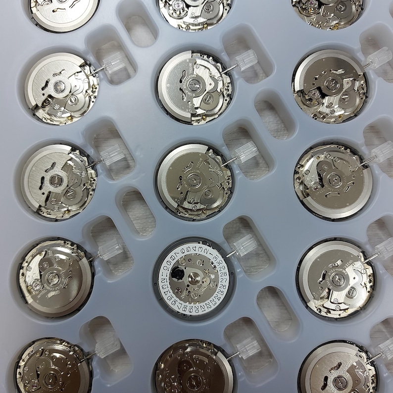 May include: A collection of watch movements, featuring intricate gears and components, arranged in a light gray plastic tray. The movements are silver-toned, showcasing the precision engineering of the watch mechanisms. Some movements have date wheels.