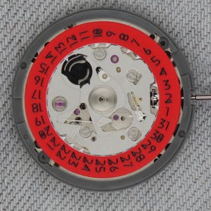 Puede incluir: Primer plano de un movimiento de reloj con un anillo exterior gris y un anillo interior rojo que muestra los días del mes en números blancos. El intrincado mecanismo presenta componentes plateados, dorados y morados.