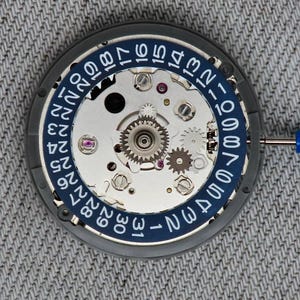 May include: Close-up of a watch movement with a blue and white date ring. The mechanical components include gears, screws, and jewels. A small blue screwdriver is in the frame. The watch movement is on a gray surface.