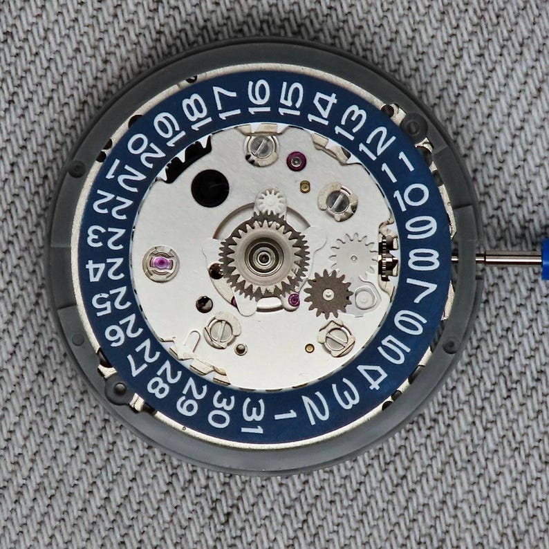 May include: Close-up of a watch movement with a blue and white date ring. The mechanical components include gears, screws, and jewels. A small blue screwdriver is in the frame. The watch movement is on a gray surface.
