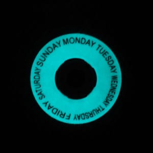 May include: A glowing teal-colored ring with the days of the week printed in white. The days of the week are arranged around the outer edge of the ring. The center of the ring is a black circle.