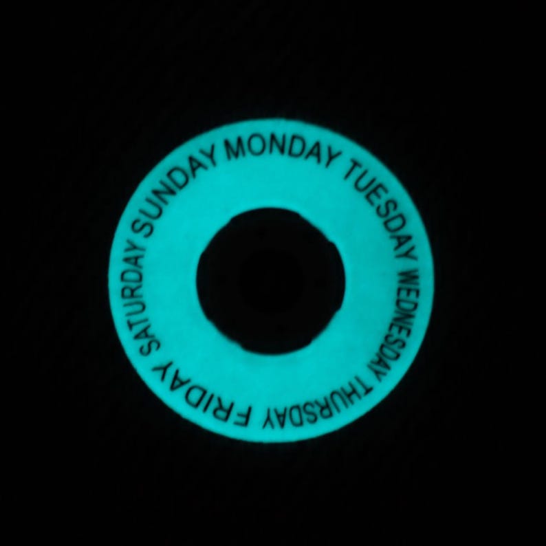 May include: A glowing teal-colored ring with the days of the week printed in white. The days of the week are arranged around the outer edge of the ring. The center of the ring is a black circle.