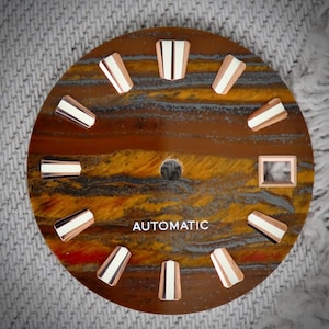 Peut inclure: Un cadran de montre avec un motif œil de tigre marron et jaune. Il comporte des index en or rose avec des accents blancs et le mot "AUTOMATIC" en blanc. Un petit carré de couleur or rose est également présent.