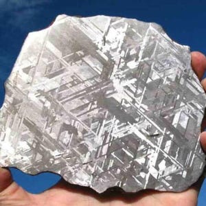 May include: A hand holds a large, irregularly shaped metallic object against a blue sky. The object has a complex, geometric pattern of intersecting lines and a silvery-gray color, suggesting a possible meteorite or geological specimen.