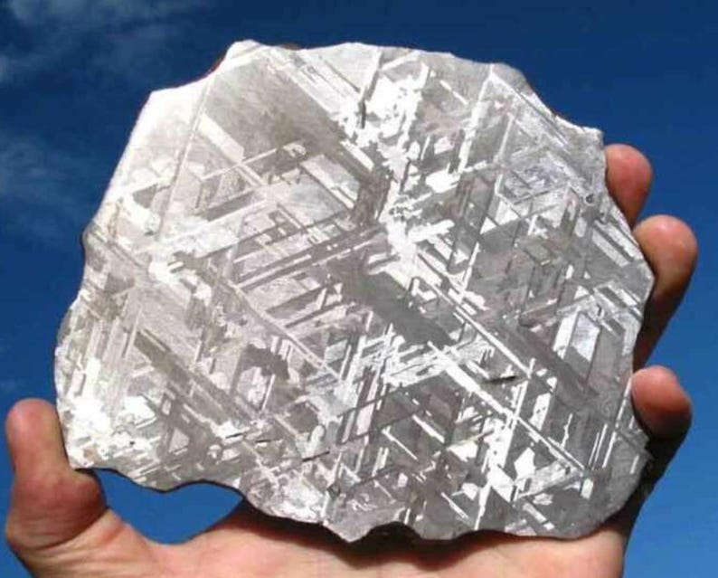 May include: A hand holds a large, irregularly shaped metallic object against a blue sky. The object has a complex, geometric pattern of intersecting lines and a silvery-gray color, suggesting a possible meteorite or geological specimen.