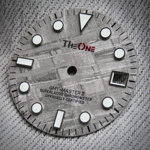 Puede incluir: Primer plano de una esfera de reloj con una superficie gris texturizada. El texto "THE ONE" está en negro y rojo. La esfera del reloj tiene marcadores circulares y rectangulares blancos, y el texto "GMT-MASTER II SUPERLATIVE CHRONOMETER OFFICIALLY CERTIFIED".