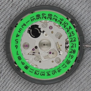 May include: Close-up of a watch movement with a green date ring and silver-colored components. The date ring displays numbers from 1 to 31. The watch mechanism is set against a gray background.