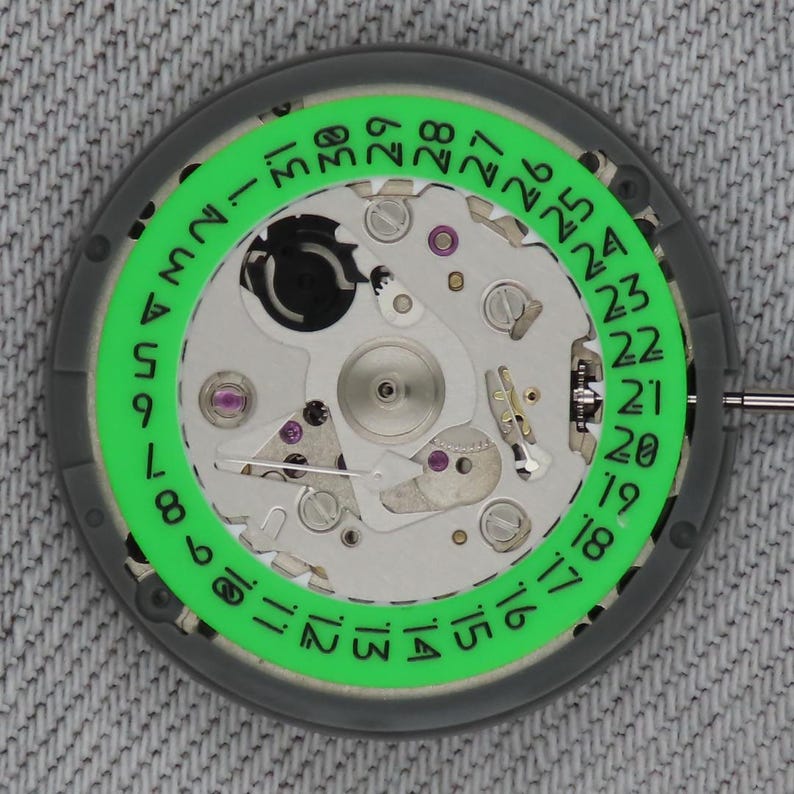 May include: Close-up of a watch movement with a green date ring and silver-colored components. The date ring displays numbers from 1 to 31. The watch mechanism is set against a gray background.