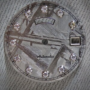 May include: Close-up of a watch face with a textured, gray metallic surface. The face features twelve diamond-like markers and the word "Automatic" in script. A rectangular window and a curved, rectangular design are also present.