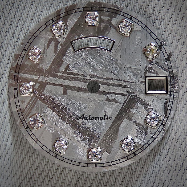 May include: Close-up of a watch face with a textured, gray metallic surface. The face features twelve diamond-like markers and the word "Automatic" in script. A rectangular window and a curved, rectangular design are also present.
