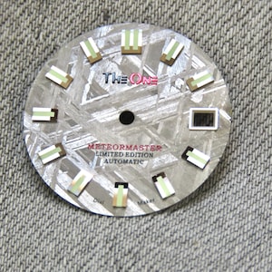 Puede incluir: Esfera de reloj circular con una superficie metálica texturizada. Cuenta con marcadores rectangulares con detalles verdes y marrones. La esfera muestra el texto "THE ONE" y "METEORMASTER LIMITED EDITION AUTOMATIC".