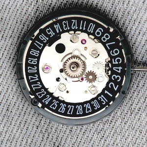 May include: Close-up of a watch movement with visible gears and a date dial. The black dial features white numbers from 1 to 31, with a silver metal casing. The intricate mechanical components are visible.