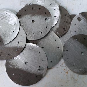 May include: A collection of circular, metallic watch faces. Each face is divided into two tones, with a lighter, brushed metal finish on one side and a darker, textured finish on the other. They have small holes and square cutouts.