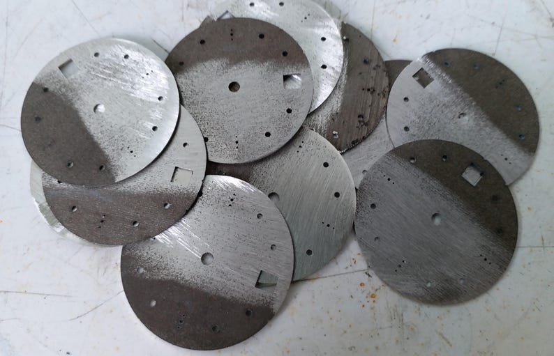 May include: A collection of circular, metallic watch faces. Each face is divided into two tones, with a lighter, brushed metal finish on one side and a darker, textured finish on the other. They have small holes and square cutouts.