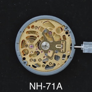 May include: A watch movement featuring detailed gold-coloured gears and mechanisms, set within a grey circular frame. The text "NH-71A" is printed below. A clear plastic stem protrudes from the side.