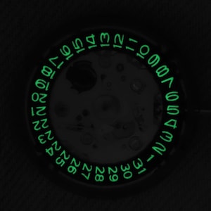 以下が含まれることがあります： 円形のデイト表示を備えた時計部品のクローズアップ。文字盤には、月の各日を示す1から31までの緑色の数字が光っています。時計のメカニズムは中央にあり、暗い背景に設定されています。