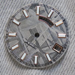 Puede incluir: Una esfera de reloj con un fondo gris texturizado y un anillo exterior negro. La esfera presenta marcadores de hora rectangulares blancos y cobrizos. La palabra "Automatic" está escrita en cursiva. La esfera del reloj tiene un diámetro de aproximadamente 3,8 cm.