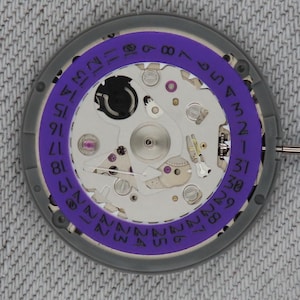 May include: Close-up of a watch movement with a purple date ring. The watch mechanism features silver and gold-colored components, including gears, screws, and jewels. The date ring is marked with numbers from 1 to 31, indicating the days of the month.