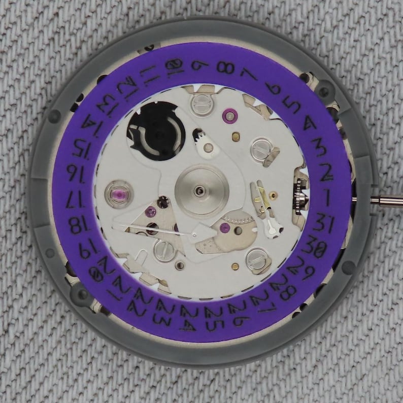 May include: Close-up of a watch movement with a purple date ring. The watch mechanism features silver and gold-colored components, including gears, screws, and jewels. The date ring is marked with numbers from 1 to 31, indicating the days of the month.