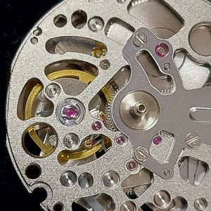 May include: Close-up of a watch movement, showcasing intricate mechanical components. The silver-colored base features numerous holes and cutouts, with gold-colored gears and ruby jewels. The design includes screws and a balance wheel, highlighting the precision of the watchmaking.