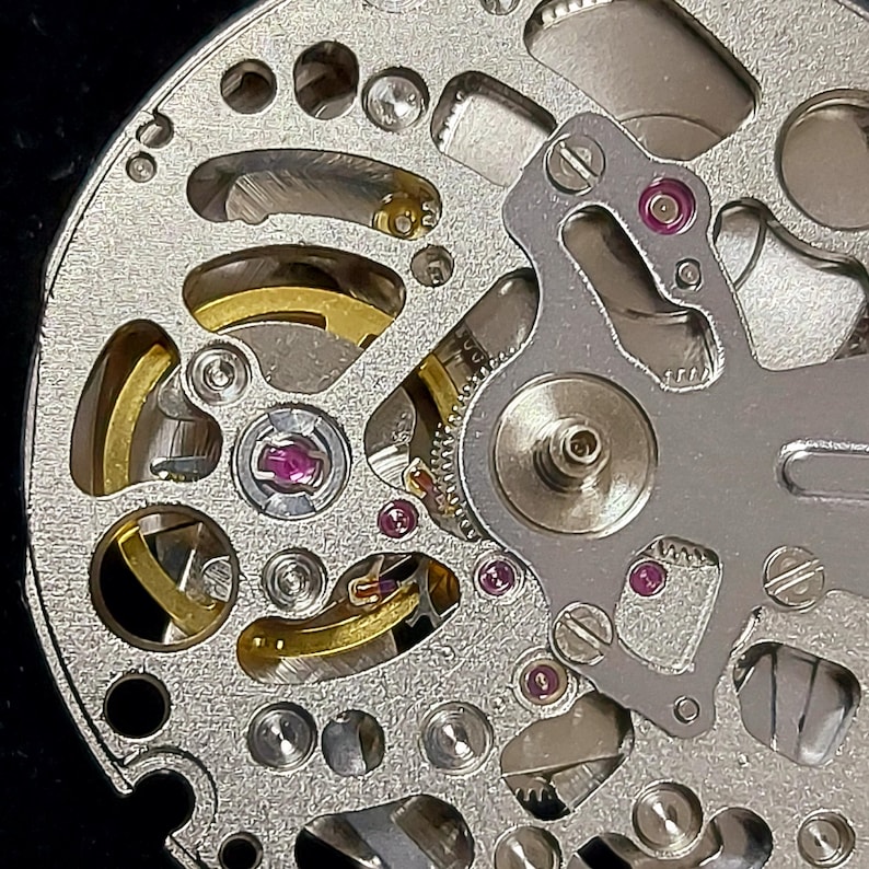 May include: Close-up of a watch movement, showcasing intricate mechanical components. The silver-colored base features numerous holes and cutouts, with gold-colored gears and ruby jewels. The design includes screws and a balance wheel, highlighting the precision of the watchmaking.