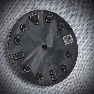 May include: A dark gray watch face with a textured pattern and silver-colored accents. The face features a date window and several circular and rectangular markers. The text "FORGED CARBON" and other details are visible.