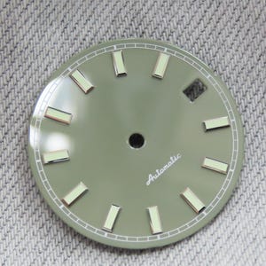 May include: A watch face with a muted green finish. It features rectangular hour markers with a silver border and a small black center hole. The word "Automatic" is written in a cursive font.