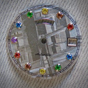 May include: A watch face with a textured, gray background and a circular design. It features a series of colorful gemstones in various colors, including green, yellow, red, and blue. The word "Automatic" is printed on the face.