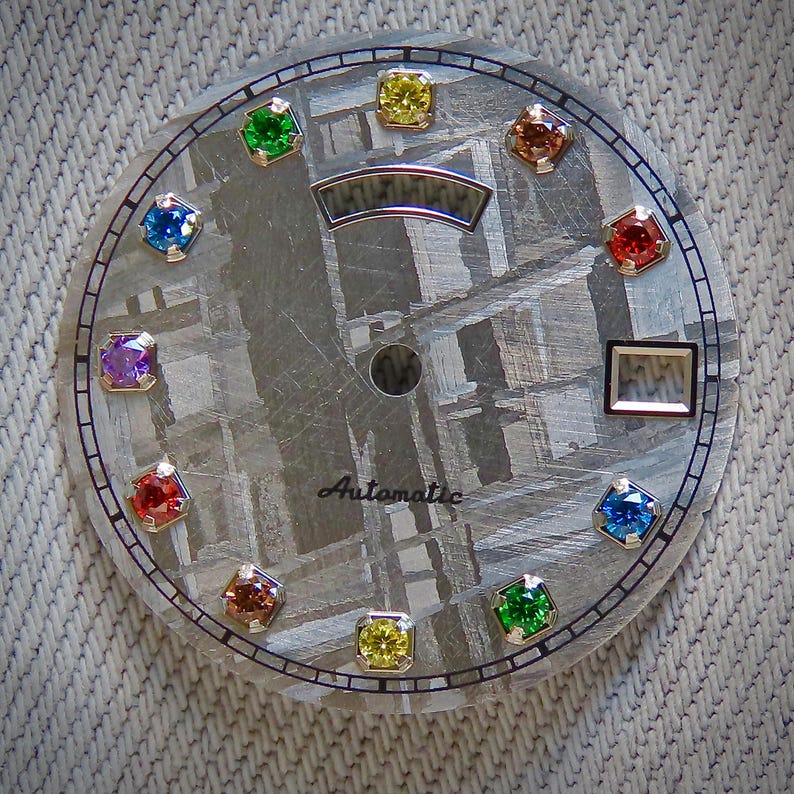 May include: A watch face with a textured, gray background and a circular design. It features a series of colorful gemstones in various colors, including green, yellow, red, and blue. The word "Automatic" is printed on the face.