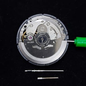 以下が含まれることがあります： 「JAPAN TWENTY-FOUR JEWELS」の文字が見える、銀色と灰色の時計ムーブメントのクローズアップ。複雑な歯車と部品が組み込まれています。緑色のドライバーが挿入されています。2つの小さな時計部品がムーブメントの下にあります。