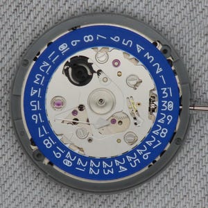 May include: Close-up of a watch movement with a blue ring displaying the days of the month. The watch mechanism is silver with small gears, jewels, and a date indicator. The outer ring is gray.