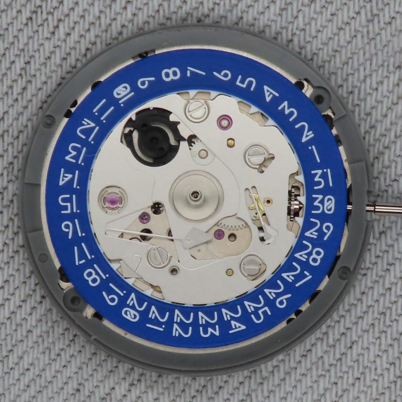 May include: Close-up of a watch movement with a blue ring displaying the days of the month. The watch mechanism is silver with small gears, jewels, and a date indicator. The outer ring is gray.