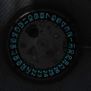 May include: Close-up of a watch face with a luminous blue date ring. The numbers 1 to 31 are visible, indicating the days of the month. The watch mechanism is partially visible in the centre.