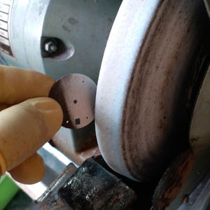 May include: A close-up of a metal disc with several small holes and a square cut-out, held near a grinding wheel. The disc is brown, and the grinding wheel is gray and brown.
