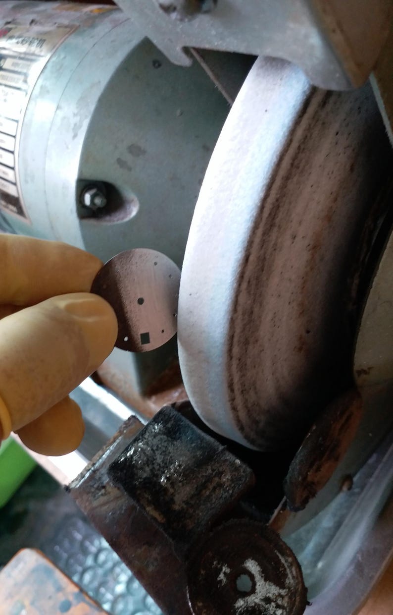 May include: A close-up of a metal disc with several small holes and a square cut-out, held near a grinding wheel. The disc is brown, and the grinding wheel is gray and brown.