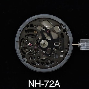 Pode incluir: Uma visão detalhada do movimento de um relógio, mostrando os complexos mecanismos internos. O movimento está alojado em uma caixa circular cinza. O texto "NH-72A" é visível na parte inferior da imagem.