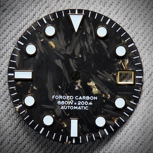 Puede incluir: Una esfera de reloj de carbono forjado negro con marcadores y agujas blancas. La esfera tiene detalles dorados y el texto "FORGED CARBON 660ft = 200m AUTOMATIC". La esfera tiene un patrón texturizado.
