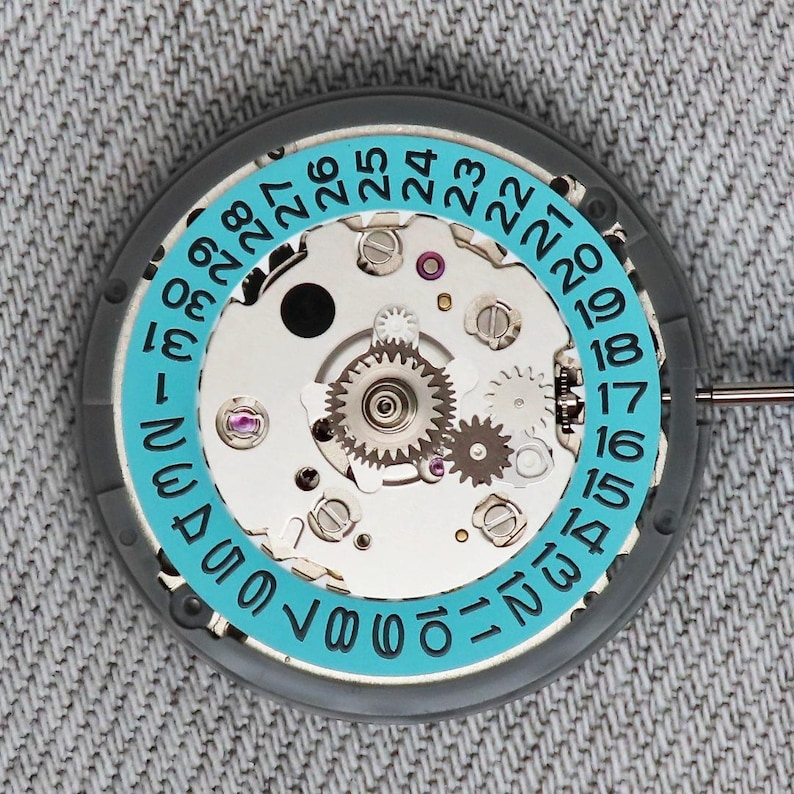 May include: A detailed view of a watch movement, featuring a gray casing and a light blue ring with numbers 1-31. The internal gears and mechanisms are visible, showcasing the intricate engineering. The watch movement is set against a gray fabric background.