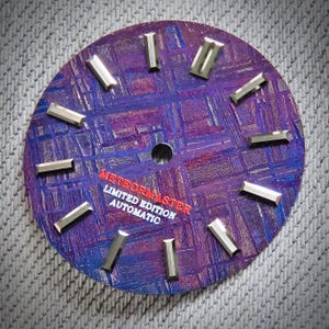 Puede incluir: Una esfera de reloj con un fondo texturizado púrpura y azul. Los marcadores de hora rectangulares plateados están espaciados uniformemente alrededor del perímetro. El texto "METEORMASTER LIMITED EDITION AUTOMATIC" está impreso en rojo y blanco.